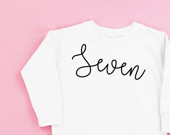 May include: A white long-sleeve sweatshirt featuring the word Seven written in black cursive script across the chest. The garment has a crew neckline, ribbed cuffs, and a ribbed hem. The text Little Mama Shirt Shop is printed in the bottom right corner.