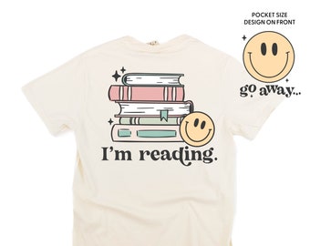 May include: A cream colored t-shirt with a stack of books and a smiley face graphic on the back. The text "I'm reading." is below the graphic. The front of the shirt has a pocket with a smiley face graphic and the text "go away..."