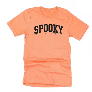 May include: A peach-coloured t-shirt featuring the word "SPOOKY" in black, dripping lettering. The shirt has a crew neck and short sleeves. The design is centred on the front, creating a Halloween theme.