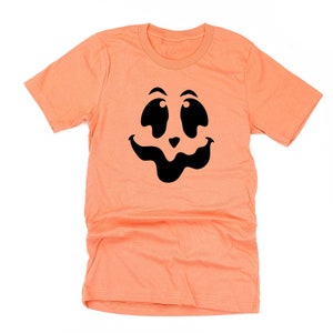 May include: A peach-colored t-shirt featuring a black jack-o'-lantern face design. The design includes large, oval eyes, a small nose, and a wide, curved mouth. The shirt is made of a soft material and has short sleeves.