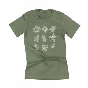 May include: Olive green t-shirt featuring a white outline design of various leaf shapes. The leaves are arranged in a grid pattern across the front of the shirt. The shirt has short sleeves and a crew neck.