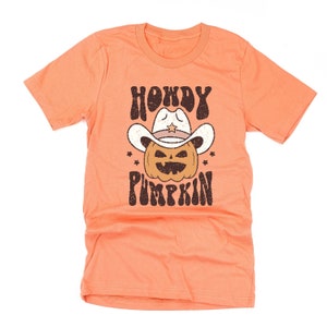 May include: Peach-colored t-shirt with a graphic design featuring a pumpkin wearing a cowboy hat. The text "HOWDY PUMPKIN" is printed above and below the pumpkin illustration. The design has a vintage, distressed look.