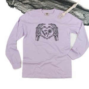 May include: A light purple long-sleeve shirt featuring a black graphic of skeleton hands forming a heart shape. Inside the heart are candy illustrations. The shirt is a comfortable, casual style.