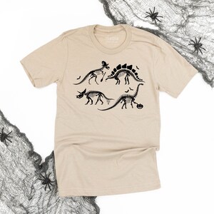 Puede incluir: Camiseta color beige con un estampado negro de esqueletos de dinosaurios decorados con motivos de Halloween. El diseño incluye un sombrero de bruja, murciélagos y una calabaza. La camiseta tiene cuello redondo y mangas cortas.