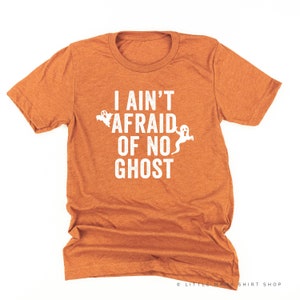 May include: Orange short-sleeved t-shirt with the phrase "I AIN'T AFRAID OF NO GHOST" in white block letters. Two white ghost illustrations are on the left and right of the text. A Halloween-themed garment.