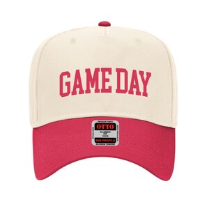 May include: A cream colored baseball cap with a red brim. The front of the cap has the text "GAME DAY" in red lettering.