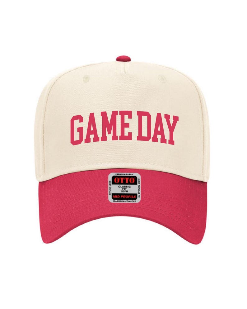 May include: A cream colored baseball cap with a red brim. The front of the cap has the text "GAME DAY" in red lettering.