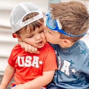 May include: Two young boys wearing red and blue shirts with the word "USA" printed on them. The boy on the left is wearing a white mesh baseball cap. The boy on the right is wearing blue goggles. The boy on the right is kissing the boy on the left on the cheek.