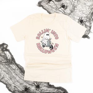 May include: Cream-colored t-shirt featuring a cartoon ghost riding a scooter. The graphic includes the text "ROLLIN' INTO HALLOWEEN" in a pink circular design. Black spiders and cobwebs are in the background, suggesting a Halloween theme.