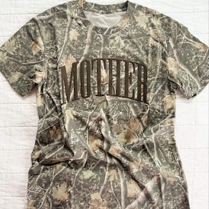 Op de afbeelding: Camouflage-patroon T-shirt met het woord "MOTHER" in bruine blokletters over de borst. Het shirt heeft een ronde hals en korte mouwen. Het camouflage-ontwerp gebruikt tinten groen, bruin en beige, waardoor een natuurlijke, outdoor esthetiek ontstaat.