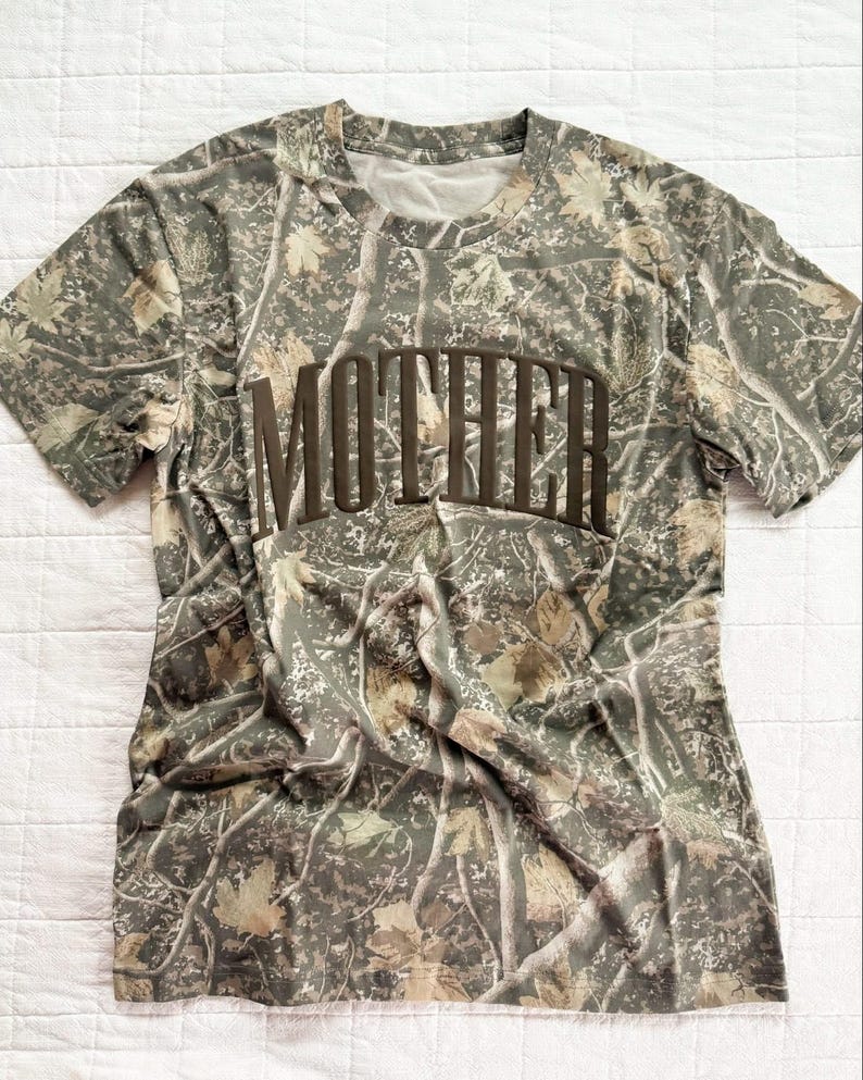 Op de afbeelding: Camouflage-patroon T-shirt met het woord "MOTHER" in bruine blokletters over de borst. Het shirt heeft een ronde hals en korte mouwen. Het camouflage-ontwerp gebruikt tinten groen, bruin en beige, waardoor een natuurlijke, outdoor esthetiek ontstaat.