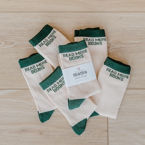 May include: A collection of cream-colored socks with green accents. Each sock features the text "READ MORE BOOKS" in green. A label with the text "LITTLE mama shirt shop" is visible on one pair of socks. The socks are arranged on a light wooden surface.