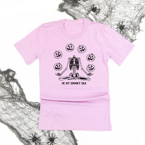 May include: A pale pink t-shirt with a black graphic of a skeleton in a yoga pose, surrounded by jack-o'-lanterns. The text "IN MY SPOOKY ERA" is printed below. Halloween-themed clothing.