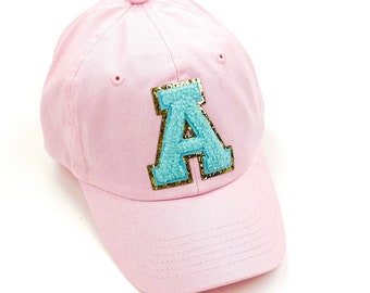 May include: A light pink baseball cap with a large, turquoise letter "A" patch. The letter is outlined in gold glitter and has a fuzzy texture. The cap is made of a soft fabric and has a curved brim.