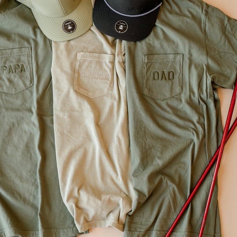 May include: Three short-sleeved shirts with front pockets, each embroidered with a different title: "PAPA", "GRANDPA", and "DAD". Two baseball caps are visible at the top. The shirts are olive green and tan.