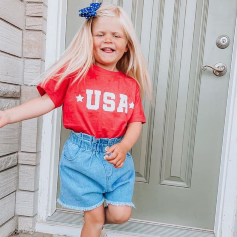 May include: A young girl wearing a red t-shirt with the word "USA" printed in white and a blue denim skirt. She is smiling and has a blue bow in her hair.