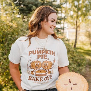 May include: Cream-colored t-shirt with a brown graphic that reads "The Great Pumpkin Pie Bake Off." The graphic features two cartoon pumpkins and a pie. The person is holding a baked pie with a golden crust.