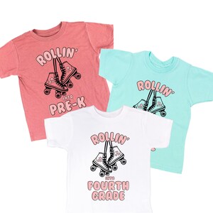 Può includere: Tre magliette a maniche corte color corallo, turchese e bianco. Ogni maglietta presenta una grafica di pattini a rotelle con la scritta "ROLLIN'" sopra e "INTO PRE-K" o "INTO FOURTH GRADE" sotto. Il design è in nero e rosa.