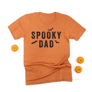 Może przedstawiać: Pomarańczowa koszulka z czarnym napisem "SPOOKY DAD" i grafiką nietoperzy. Obraz zawiera małe pomarańczowe dynie, sugerując motyw Halloween. Koszulka wykonana jest z miękkiego materiału.