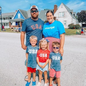 May include: A family of five wearing red, white, and blue clothing with the word "USA" on their shirts. The family is standing in front of a white house with a blue sky in the background.