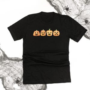 May include: Black t-shirt featuring four watercolor-style jack-o'-lanterns with different expressions. The pumpkins are orange with green stems and carved faces. The shirt is a crew neck and made of a soft material. Halloween themed apparel.