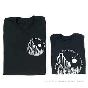 May include: Two black t-shirts with a white mountain and forest design. The larger shirt says "Explore More" and the smaller shirt says "Exploring with Dad".