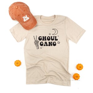 May include: A beige t-shirt with the words "GHOUL GANG" in black, a skeleton hand peace sign, a crescent moon, and stars. An orange baseball cap with "Fall Babe" is next to the shirt. Small orange pumpkins are scattered around the shirt.