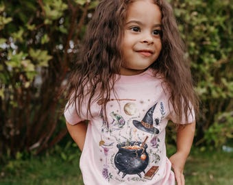 May include: A light pink t-shirt featuring a whimsical witch-themed design. The graphic includes a black cauldron, a witch's hat, a frog, a moon, and other magical elements. The shirt is worn by a young person.