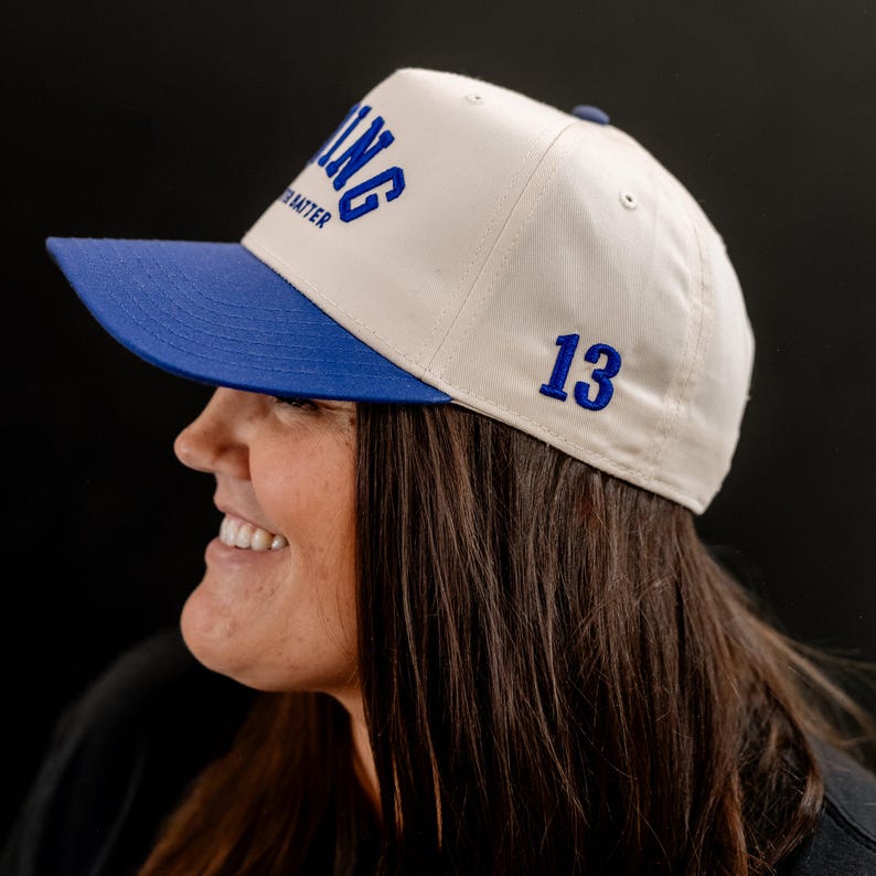 May include: A white baseball cap with a blue brim and the word "LONG" embroidered on the front. The number "13" is embroidered on the back of the cap.