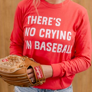 Puede incluir: Una camiseta roja de manga larga con el texto "THERE'S NO CRYING IN BASEBALL" en blanco. Una persona sostiene un guante de béisbol marrón.