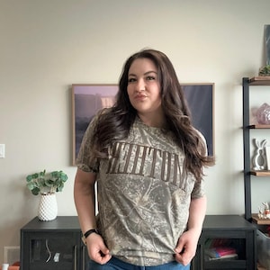 Op de afbeelding: Een t-shirt met korte mouwen en camouflagepatroon met het woord "KEENUM" in bruine letters op de borst. Het shirt wordt gecombineerd met een blauwe spijkerbroek. De persoon draagt een zwart horloge.
