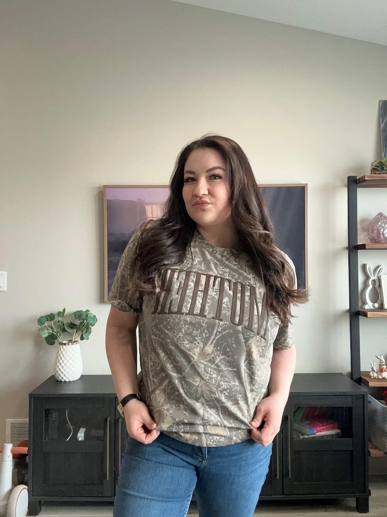 Op de afbeelding: Een t-shirt met korte mouwen en camouflagepatroon met het woord "KEENUM" in bruine letters op de borst. Het shirt wordt gecombineerd met een blauwe spijkerbroek. De persoon draagt een zwart horloge.