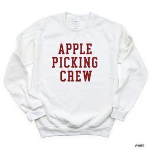 May include: White crewneck sweatshirt with the text "APPLE PICKING CREW" in a bold, maroon font. The sweatshirt is a solid white color and has a classic, casual style. The image is a close-up of the front of the sweatshirt.