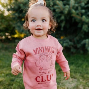 May include: A child's dusty rose-coloured sweatshirt with the words "MINI MUG CLUB" in red, and a graphic of a mug with cinnamon sticks and star anise. The words "REFILLS ENCOURAGED" are also printed on the sweatshirt. The child is smiling and looking at the camera.