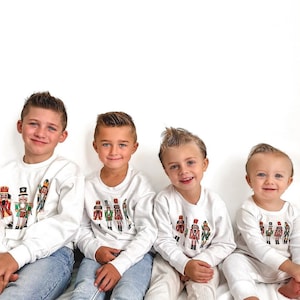 May include: Four children wearing white sweatshirts and pajamas with a colorful nutcracker design. The children are smiling and sitting together. The sweatshirts have long sleeves and a crew neck.