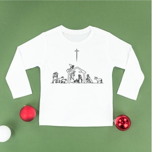 Puede incluir: Camiseta blanca de manga larga con un dibujo lineal negro de un pesebre. El diseño incluye figuras de María, José, el niño Jesús, ángeles y animales bajo una estrella. Adornos navideños rojos y blancos sobre un fondo verde.