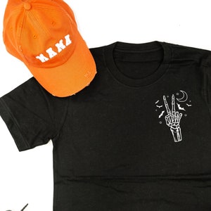 Könnte beinhalten: Schwarzes T-Shirt mit einer weißen Skeletthand, die ein Friedenszeichen zeigt, Fledermäusen, Sternen und einem Mond. Eine orangefarbene Baseballkappe mit dem Wort "NANA" in weißer Schrift liegt neben dem Shirt.