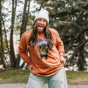 May include: Orange long-sleeve shirt with a graphic of a tongue with fangs and the color green. The person in the image is wearing a white beanie and light blue jeans. The background is a blurred outdoor scene with trees.
