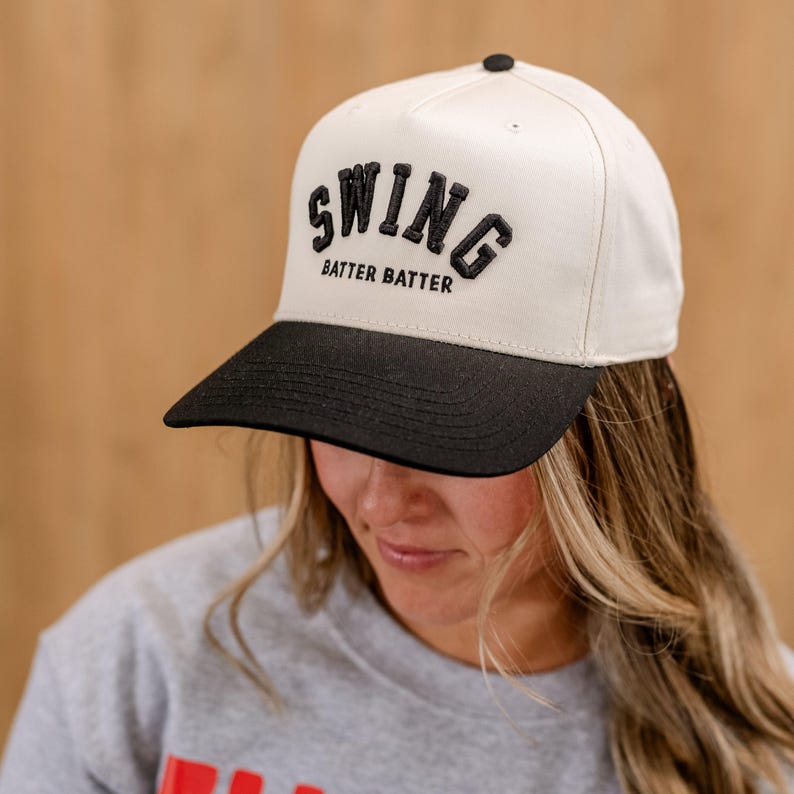 May include: A white and black baseball cap with the text "SWING BATTER BATTER" embroidered on the front.