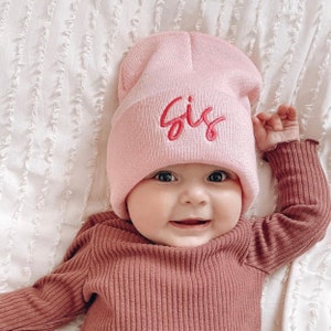 Puede incluir: Un gorro de punto rosa con la palabra "Sis" bordada en rojo en la parte delantera. El gorro lo lleva puesto un bebé con un atuendo rosa y marrón.