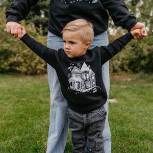 May include: Black sweatshirt with a white haunted house graphic and the text "ENTER IF YOU DARE". The child is wearing gray pants. The adult is wearing a black sweatshirt and blue jeans. The background is green grass.