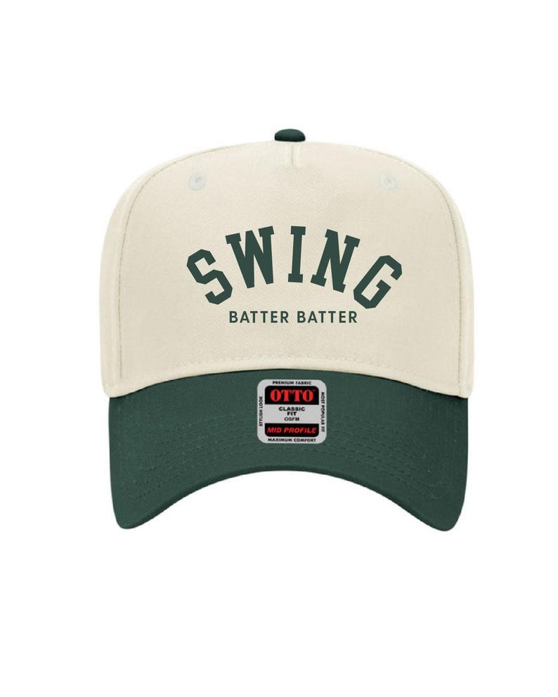 May include: A cream colored baseball cap with a green brim. The front of the cap has the word "SWING" in green letters with the words "BATTER BATTER" underneath. The back of the cap has a black and red label with the word "OTTO" in red letters.