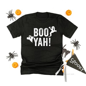 May include: Black t-shirt with the words "BOO YAH!" in white, along with ghost graphics. The shirt is surrounded by Halloween decorations, including spiders, pumpkins, and a "Spooky" flag.