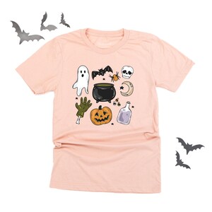 May include: A light pink t-shirt featuring Halloween-themed illustrations. The design includes a ghost, bat, spider, skull, cauldron, pumpkin, potion bottle, and a zombie hand. The shirt is a crew neck style.