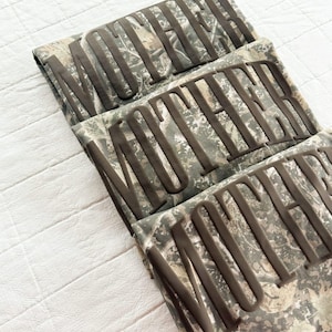 Op de afbeelding: Drie gevouwen shirts met camouflagepatroon en het woord "MOTHER" in verhoogde, donkerbruine letters. De shirts zijn netjes op een wit oppervlak gevouwen, met een camouflageontwerp in groen, bruin en grijs.