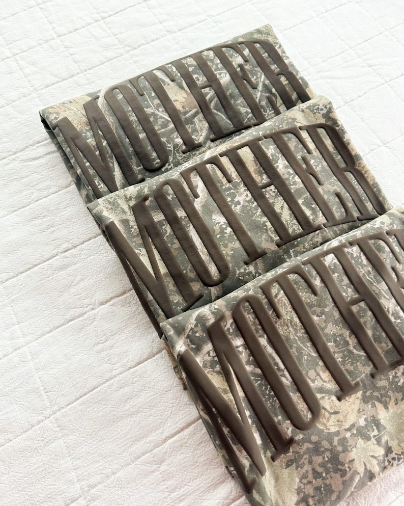 Op de afbeelding: Drie gevouwen shirts met camouflagepatroon en het woord "MOTHER" in verhoogde, donkerbruine letters. De shirts zijn netjes op een wit oppervlak gevouwen, met een camouflageontwerp in groen, bruin en grijs.