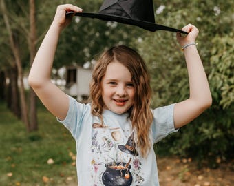 May include: A black witch hat held up over a light blue t-shirt with a witchy graphic. The graphic includes a cauldron, witch hat, and other magical elements. The image is taken outdoors with a natural background.