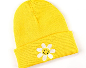Puede incluir: Un gorro amarillo con una margarita bordada en blanco con una cara sonriente en el centro.
