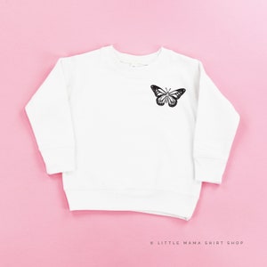 Pu&ograve; includere: Felpa bianca a maniche lunghe con una grafica di farfalla nera sul davanti. Il testo "LITTLE MAMA SHIRT SHOP" &egrave; stampato in nero nella parte inferiore dell'immagine.