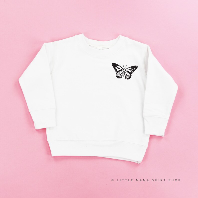 Pu&ograve; includere: Felpa bianca a maniche lunghe con una grafica di farfalla nera sul davanti. Il testo "LITTLE MAMA SHIRT SHOP" &egrave; stampato in nero nella parte inferiore dell'immagine.